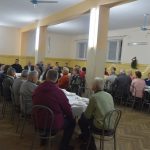 Grupa osób siedzących przy stołach na rozlelglej sali. Na stale znajduje się zastawa.