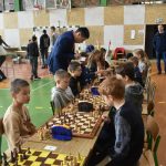 Grupa młodych szachistów na sali gimnastycznej w trakcie rozgrywek. W te widać grupę dorosłych i młodzieży.