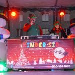 Kobieta i mężczyzna stojący na scenie w stroju świątecznych elfów. W tle dwa dmuchane bałwany i gmach bloku mieszkalnego.