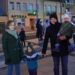 Grupa osób w strojach zimowych, różnej płci i wieku, zebrana na miejskim rynku, W tle budynek apteki.