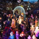 Grupa osób różnej płci i wieku zebrana na rynku. W tle widoczne są instalacje świąteczne.