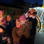 Grupa dzieci dorosłych i dzieci zebrana na rynku. W tle świeci się masywna instalacja świąteczna.