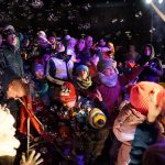 Grupa osób różnej płci i wieku zebrana na rynku. Panuje noc. Wszyscy ubrani są w ciepłe, zimowe stroje.