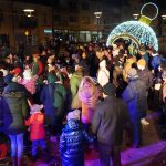 Grupa osób różnej płci i wieku zebrana na rynku. Panuje noc. Wszyscy ubrani są w ciepłe, zimowe stroje.