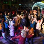 Grupa osób różnej płci i wieku zebrana na rynku. Panuje noc. Wszyscy ubrani są w ciepłe, zimowe stroje.