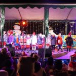 Grupa dzieci wystepująca na scenie na otwartej przestrzeni. Panuje noc. Wszyscy ubrani są w zimowe stroje.