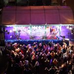 Grupa osób różnej płci i wieku zebrana na rynku w zimowa noc i spoglądająca na oświetloną scenę.