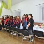 Grupa młodzieży, głównie w strojach sportowych stojąca przy długim stole na rozległej sali.