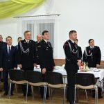 Grupa dorosłych osób, głownie mężczyzn stojąca przy długim stole. Osoby te ubrane są w zdecydowanej większości w mundury i garnitury.