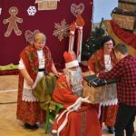 Osoba w stroju Mikołaja wręczająca prezent młodemu chłopakowi. Obok stoją dwie kobiety w strojach ludowych.