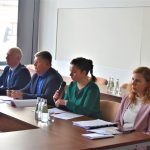 Dwie kobiety i dwóch mężczyzn zasiadający za rozległym biurkiem na dużej sali. Jedna z kobiet przemawia do mikrofonu.