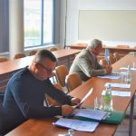 Trzech dorosłych mężczyzn zasiadająca za rozległym biurkiem. Przy nich lezą dokumenty i woda.