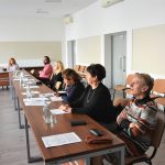 Grupa dorosłych kobiet i mężczyn zasiadająca za rozległym biurkiem. Przy nich lezą dokumenty i woda.