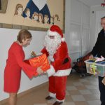 Mężczyzna przebrany w strój mikołaja, i mężczyzna ubrany w ciemny płaszcz wręczają prezent opakowany w czerwony papier kobiecie o krótkich włosach w czerwonej sukience. Znajdują się na korytarzu.