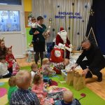 Mężczyzna w ciemnym płaszczu wręcza dzieciom, siedzącym na dywanie w przedszkolu pudełko klocków. Z boku siedzi mężczyzna przebrany w strój mikołaja.