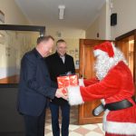 Męzczyzna w stroju mikołaja wręcza mężczynie w garniturze prezent zapokowany z czerwony papier. Za nimi stoi mężczyzna w ciemnym płaszczu i się uśmiecha.