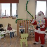 Mężczyzna w stroju mikołaja wręcza pudełko klocków dzieciom, siedzącym w fotelikach do karmienia.