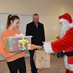 Mężczyzna w stroju mikołaja wręcza pudełko klocków i brązowe reklamówki kobiecie w pomarańczowym swetrze. Za nimi stoi mężczyzna w ciemnym płaszczu i się uśmiecha,.