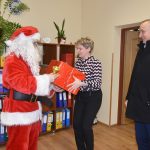 Mężczyzna w stroju mikołaja wręcza prezent opakowany w czerwony papier kobiecie. Za nimi mężczyzna w ciemnym garniturze się uśmiecha.