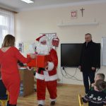 Mężczyzna w stroju mikołaja wręcza prezent opakowany w czerwony papier kobiecie. Za nimi mężczyzna w ciemnym garniturze się uśmiecha.