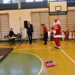 Grupa osób na rozległej sali. Na pierwszym planie widać mężczyznę przebranego za Mikołaja, Mężczyznę w garniturze, oraz kobietę w czerwonym swetrze. Obok stoi grupa dzieci.