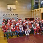 Grupa osób głownie dzieci siedząca na ławkach i krzesełkach w sali gimnastycznej w mikołajkowych strojach.