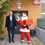 Mężczyzna w stroju mikołaja wraz z drugim mężczyzną w ciemnym stroju stoją przed budynkiem szkoły.