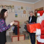 Mężczyzna w stroju Mikołaja oraz mężczyzna w jasnej koszuli i ciemnym płaszczu wręczają prezent kobiecie.