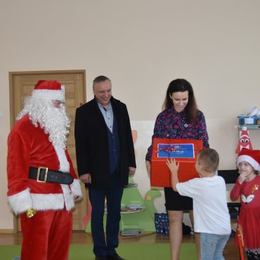 Grupa osób stoi w sali przedszkolnej. Mężczyzna w stroju mikołaja, kobieta w kolorowej bluzce wręcza prezent opokowany w czerwony papier chłopcu w białej koszulce.