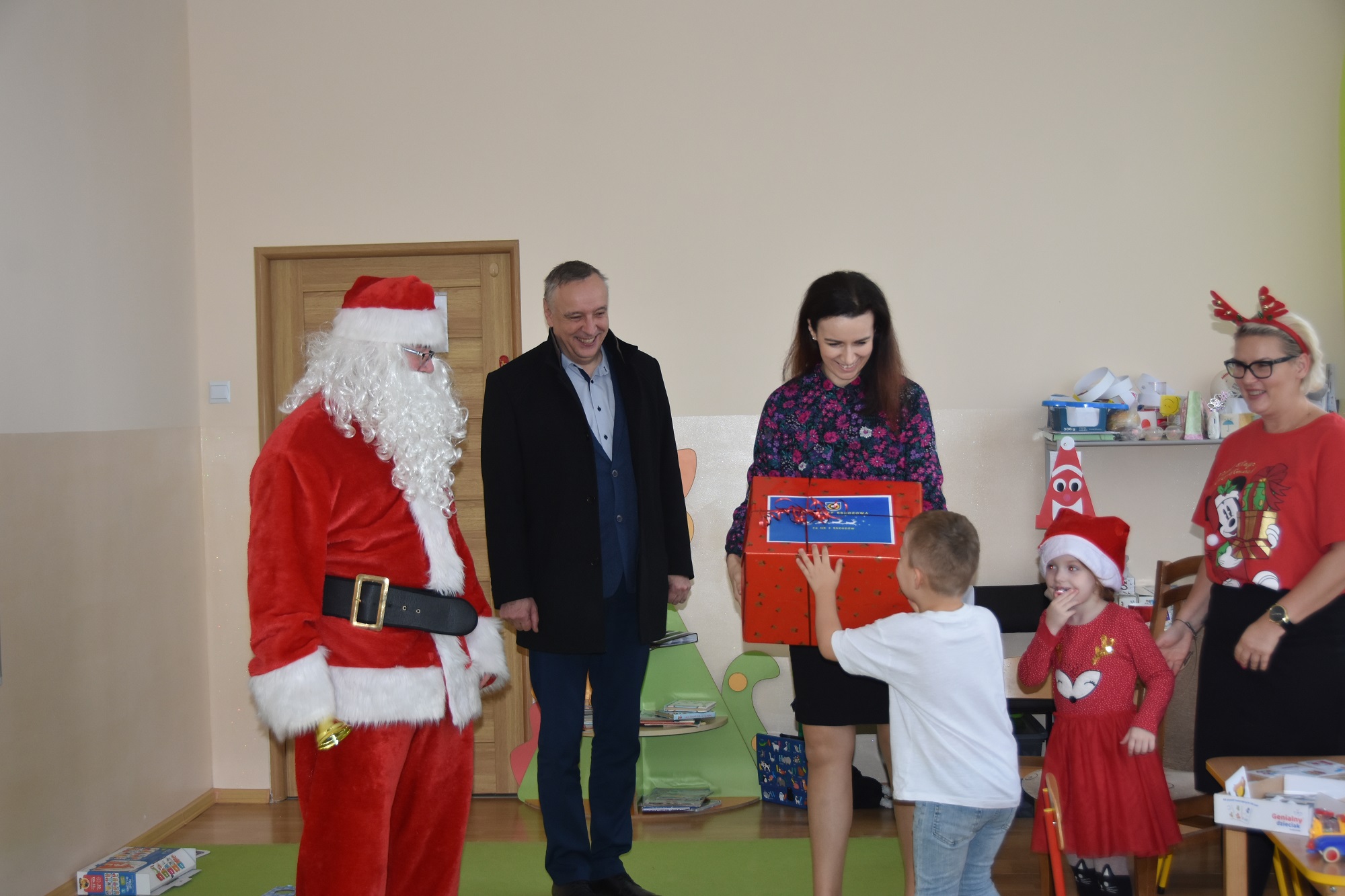 Grupa osób stoi w sali przedszkolnej. Mężczyzna w stroju mikołaja, kobieta w kolorowej bluzce wręcza prezent opokowany w czerwony papier chłopcu w białej koszulce.