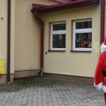 Mężczyzna w stroju Mikołaja macha do dzieci za oknem. W ręce trzyma dzwonek.