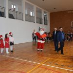Mężczyzna w stroju Mikołaja na sali gimnastycznej. W tle grupa dzieci i dwóch mężczyzn.