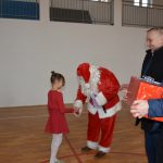 Osoba w stroju Mikołaja otrzymuje prezent od dziewczynki w czerwonej sukience. Obok stoi dwóch mężczyzn jeden trzyma prezent.