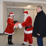 Mężczyzna w ciemnym płaszczu i mężczyzna w stroju Mikołaja, przekazujący prezent kobiecie w czerwonym stroku.