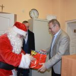 Mężczyzna w stroju Mikołajka przekazuje prezent mężczyźnie w jasnym garniturze W tle mężczyzna w ciemnym płaszczu.