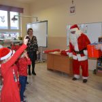 Męzczyzna w stroju mikołaja wita się z dziećmi w sali lekcyjnej. Obok stoją meźczyźni w garniturach i kobieta w spódnicy.