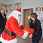 Mężczyzna w stroju mikołaja przekazuje kobiecie o krótkich włosach prezent opokowany w czerwony papier ozdobny. Za nimi usmiecha się mężczyzna w ciemnym stroju.