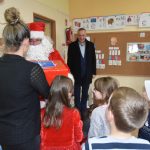 Mężczyzna w stroju mikołaja przekazuje prezent na ręce kobiety ubranej w czarną koszulkę. Przed nimi stoją dzieci i mężczyzna w ciemnym płaszczu.