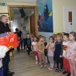 Mężczyzna w stroju mikołaja i mężczyzna w ciemnym płaszczu witają się z grupą dzieci w sali przedszkolnej.