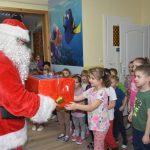 Mężczyzna w stroju mikołaja przekazuje na ręce dzieci prezent opokowany w czerowny papier.