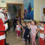 Mężczyzna w stroju mikołaja i mężczyzna w ciemnym płaszczu witają się z grupą dzieci w sali przedszkolnej.