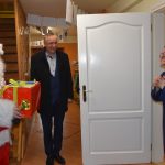 Mężczyzna przebrany w czerowny stroj mikołaja wraz z mężczyzną w ciemnym płaszczu stoją w pomiesczeniu i witaja się z kobietą ubraną w granatowy habit.