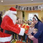 Mężczyzna w stroju mikołaja przekazuje na ręce dziewczynki duży prezent zapokowany w czerowny papier. Obok nich stoi zakonnica ubrana w granatowy habit.