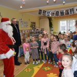 Mężczyzna w stroju Mikołaja i mężczyzna w czarnym płaszczu spotykają się z grupą dzieci w sali przedszkolnej. Obok nich stoi siostra zakonna ubrana w granatowy habit.