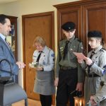 Dorosły mężczyzna w garniturze stoi z grupą dzieci i dorosłą kobieta w strojach harcerskich. Jedna z dziewczynek czyta z kartki.