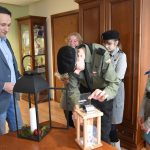 Dorosły mężczyzna w garniturze stoi z grupą dzieci i dorosłą kobieta w strojach harcerskich. Chłopiec zapala świece w dużym szklanym lampionie.