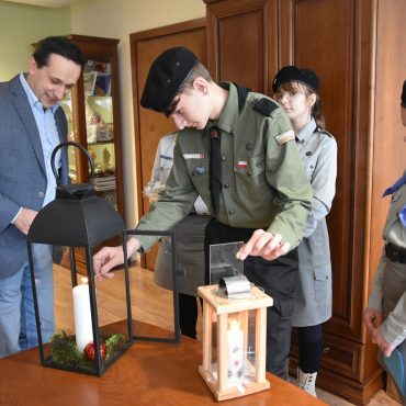 Dorosły mężczyzna w garniturze stoi z grupą dzieci i dorosłą kobieta w strojach harcerskich. Chłopiec zapala świece w dużym szklanym lampionie.