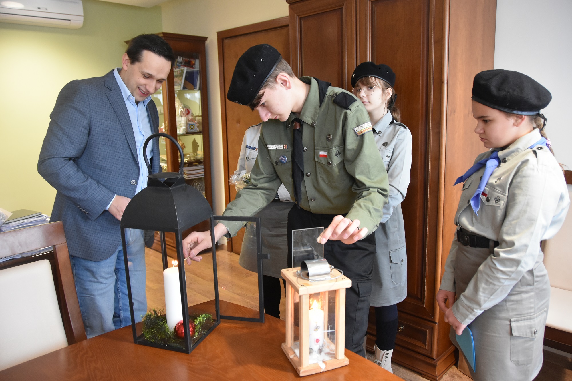 Dorosły mężczyzna w garniturze stoi z grupą dzieci i dorosłą kobieta w strojach harcerskich. Chłopiec zapala świece w dużym szklanym lampionie.