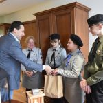 Dorosły mężczyzna w garniturze przekazuje grupie harcerzy wraz z opiekunką upominki.