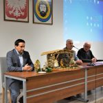 Trzech dorosłych mężczyzn w garniturach siedzących za prezydium na dużej sali. Na biurku znajduje się szopka.